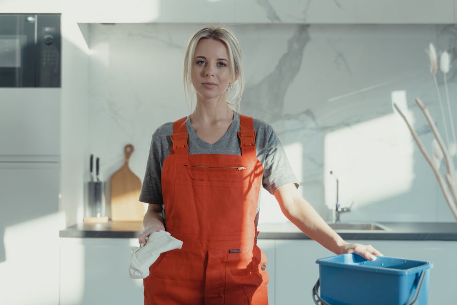 A professional cleaner wearing a red apron and holding a cleaning cloth stands in a modern kitchen with white marble walls and stainless steel appliances. The countertop is empty, clean, and reflectiv