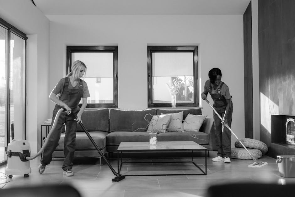 Two professional cleaners from Cleaners Elephant and Castle are performing surface cleaning in a modern, well-lit living room. One cleaner is using a vacuum cleaner to clean the wooden floor near a so